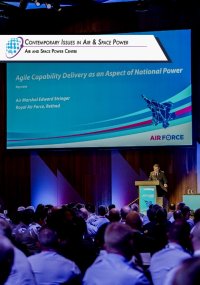 E. Stringer on stage at Chief of Air Force Symposium delivering keynote speech. Large screen in the background displaying powerpoint with title of speech.
