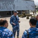 Commanding Officer Officers Training School, Wing Commander Garth Herriot giving a tour to Indonesian Air Force members in a circle, standing in front of a grey display board on the side of a building