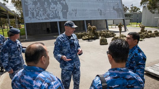 Commanding Officer Officers Training School, Wing Commander Garth Herriot giving a tour to Indonesian Air Force members in a circle, standing in front of a grey display board on the side of a building